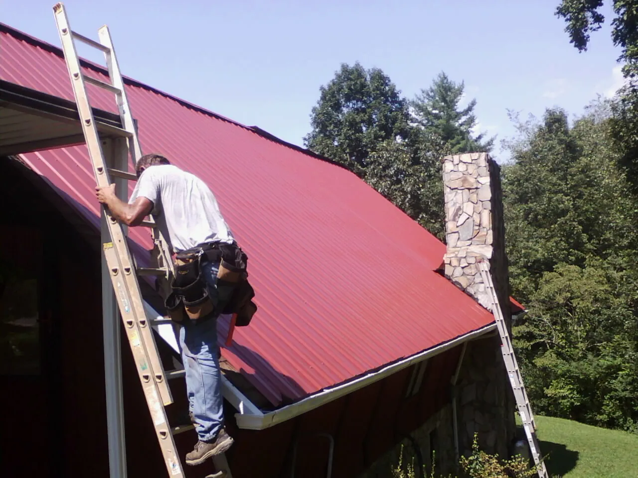 Roofer on a ladder beside a red metal roof and stone chimney — Soffit & Fascia Repair by Roofing Brothers in Long Creek